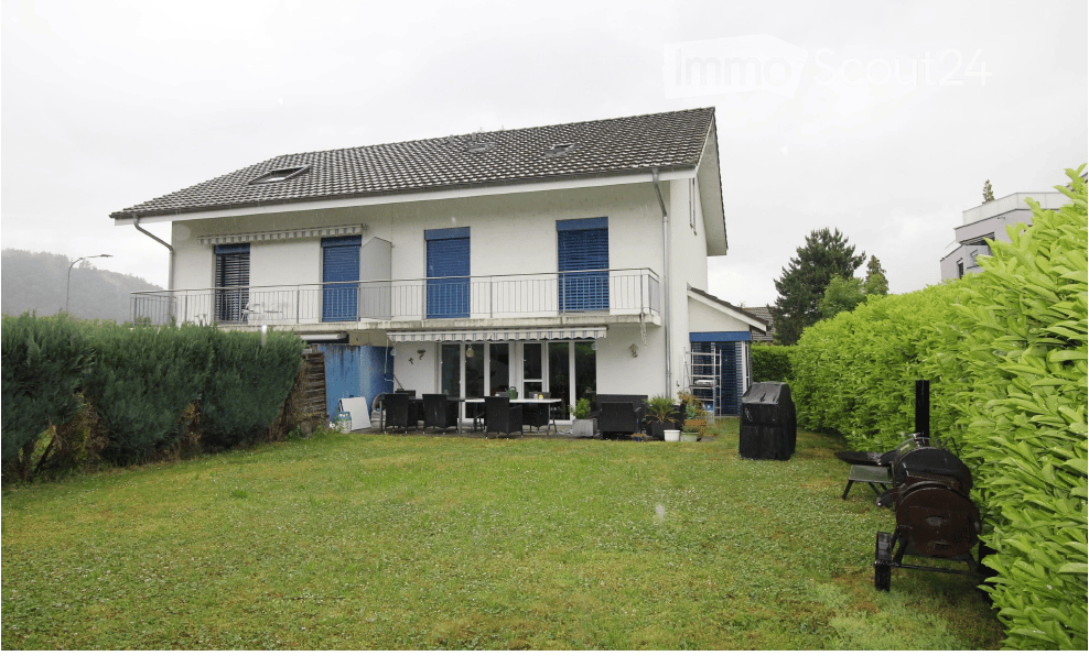 Two-story white house with blue shutters, a balcony, and a well-maintained lawn with outdoor furniture and a barbecue.