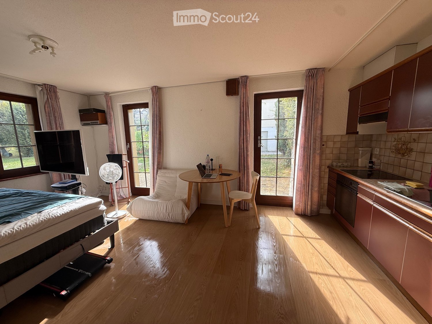 A well-lit room with a bed, a chair, a small table with a laptop, a fan, a television, and a kitchen area with cabinets and a dishwasher.
