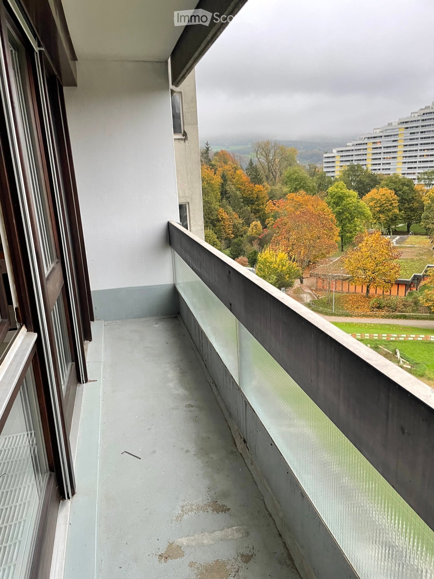 Empty balcony with concrete floor, glass railing, and view of trees and buildings.