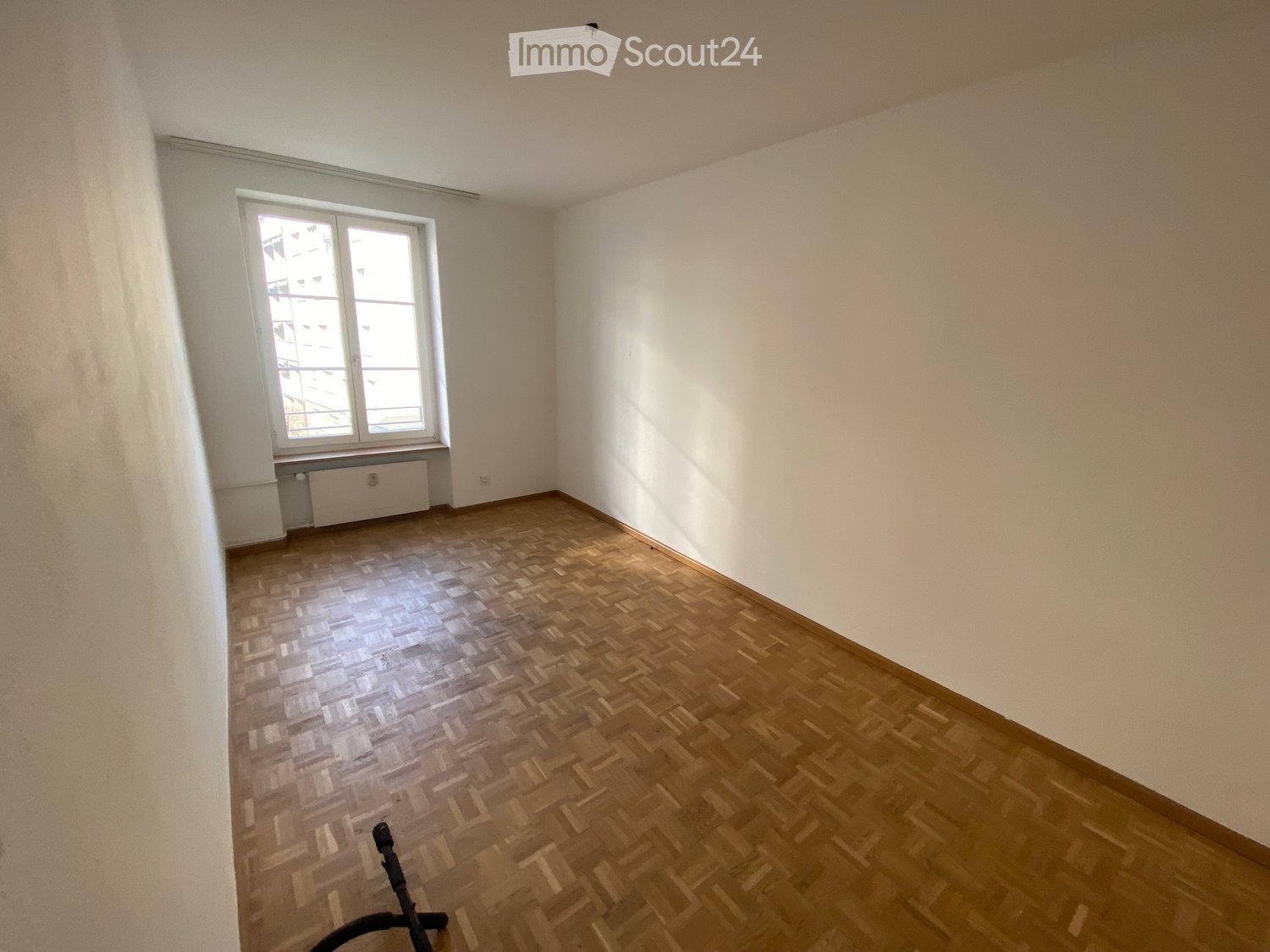 Empty room with wooden herringbone flooring, white walls, and a window with a radiator below it.
