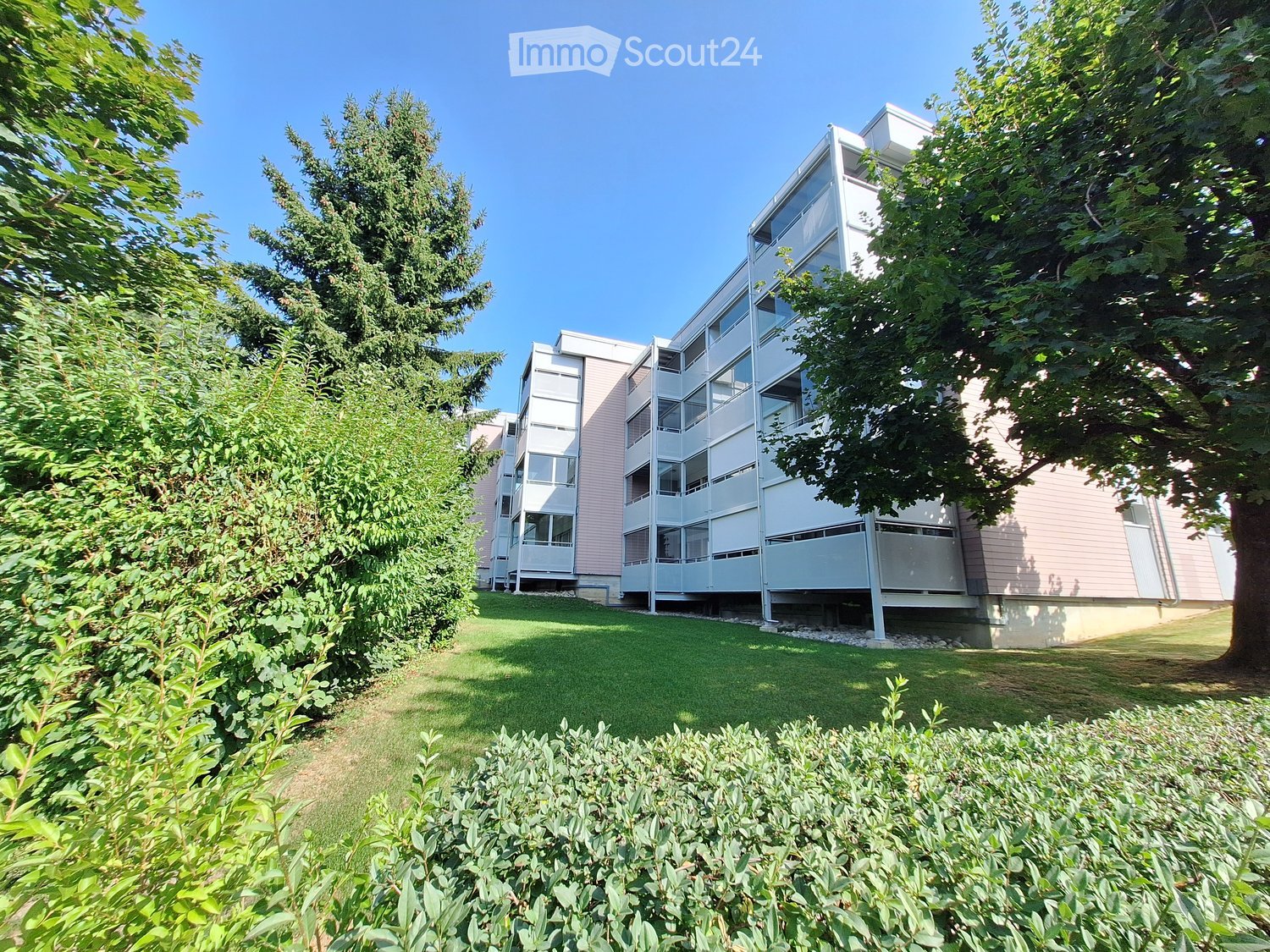Modern multi-story residential building with white and pink exterior, surrounded by green lawn and trees.