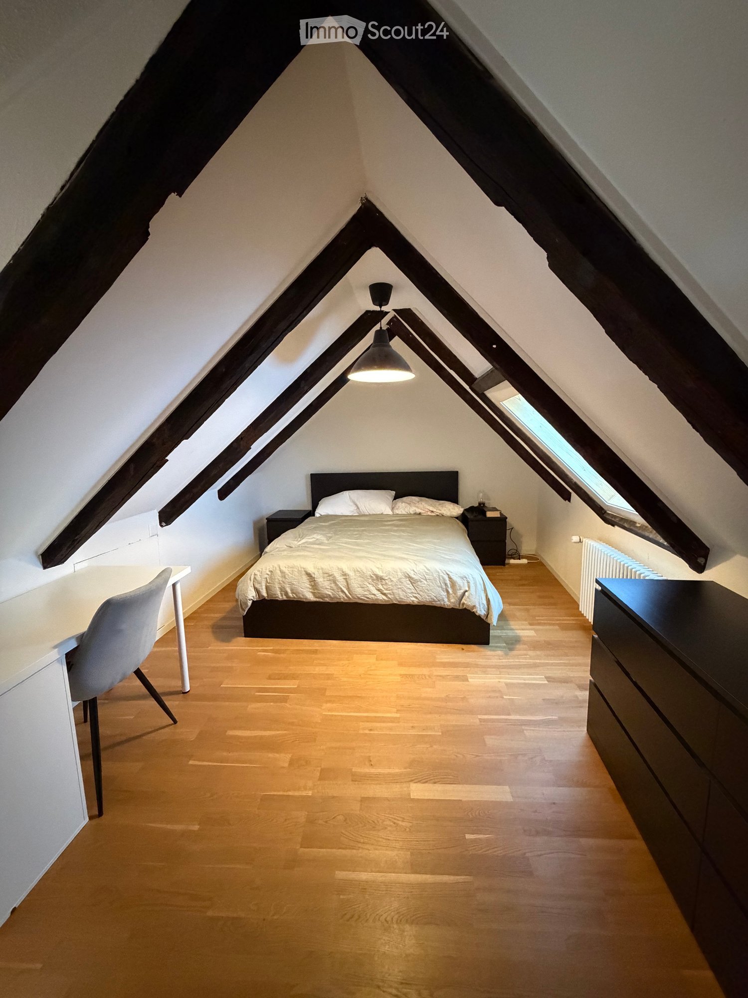 Cozy attic bedroom with wooden floors, slanted ceilings, and large windows. Features a bed with white bedding, a nightstand, a desk with a chair, and a hanging pendant light.