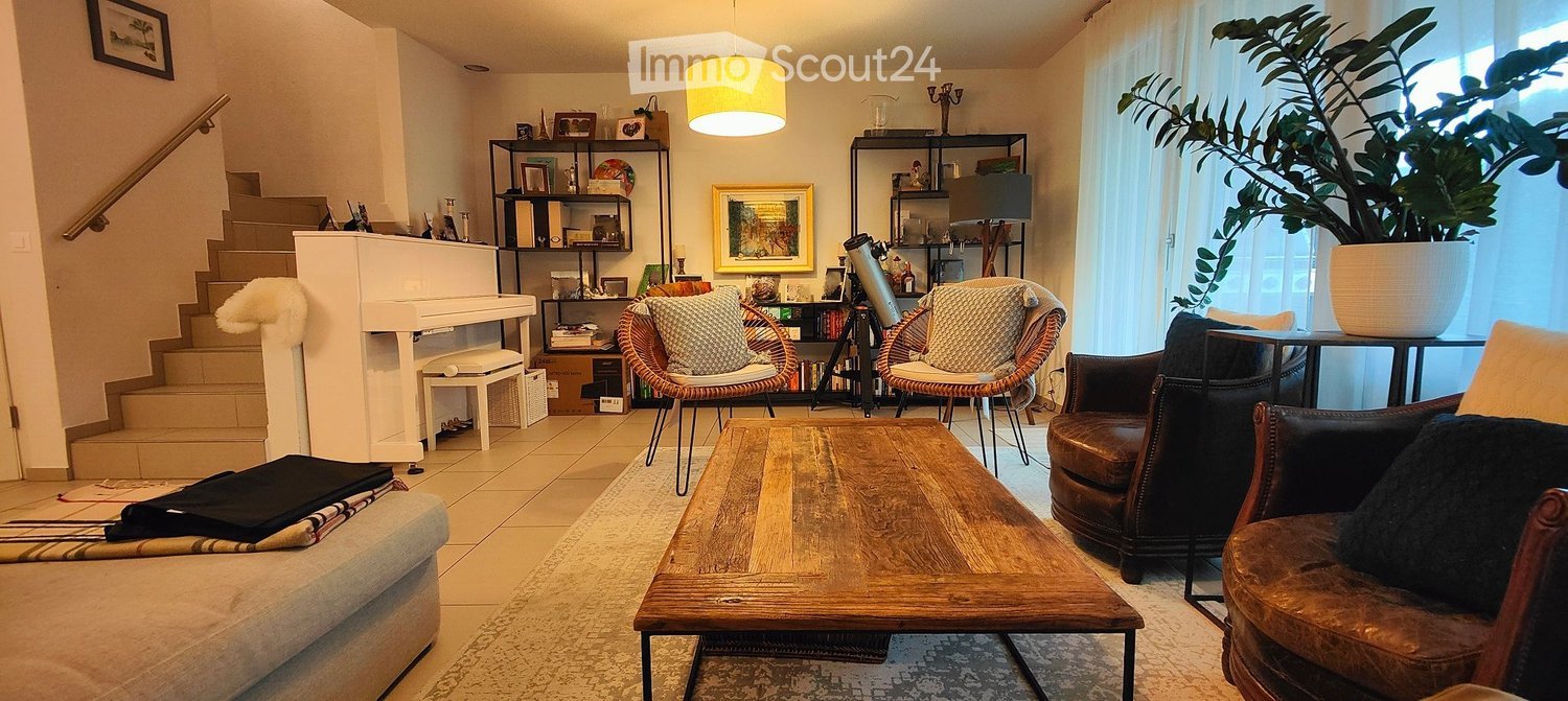 A cozy living room with a wooden coffee table, leather sofa, wicker chairs, a small piano, and a large potted plant near the window. Shelves hold books and decorative items, and a staircase leads upwards.