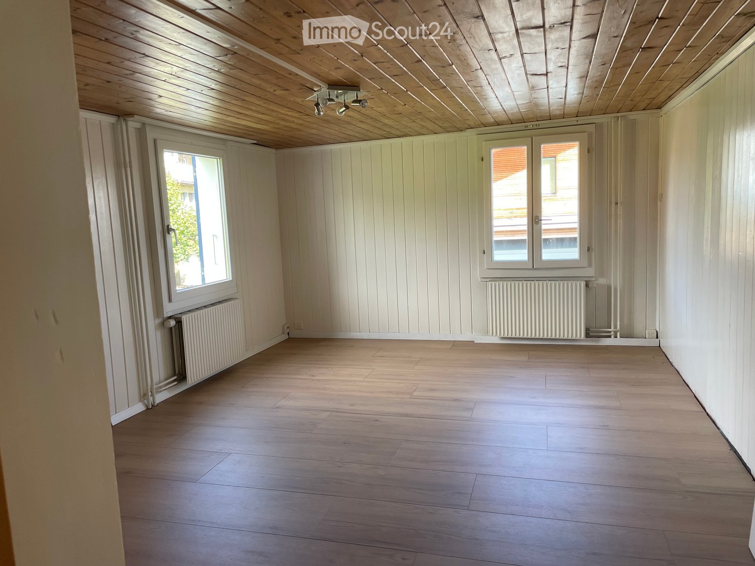 Empty room with wooden flooring, white paneled walls, and large windows with radiators beneath them.