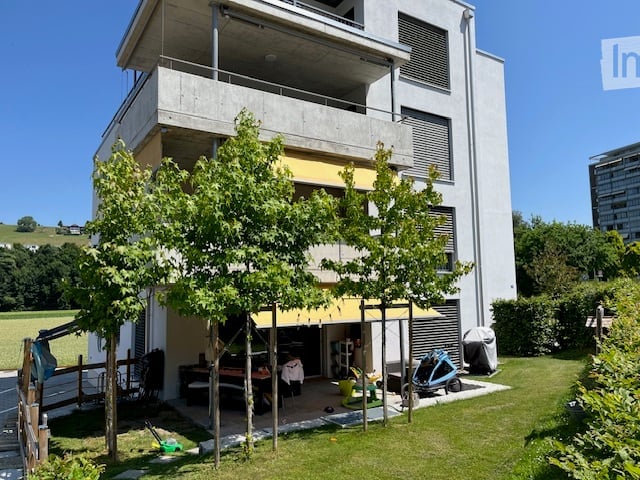 Modern two-story building with a balcony, surrounded by greenery and a small outdoor seating area.