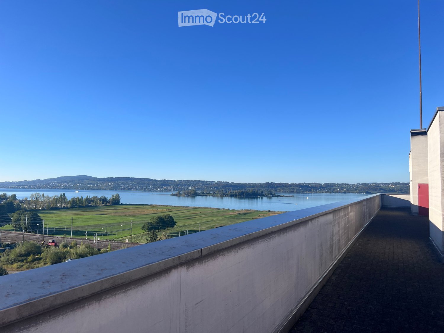 Balcony with a low concrete railing, overlooking a scenic view of a lake, distant hills, and a green field.