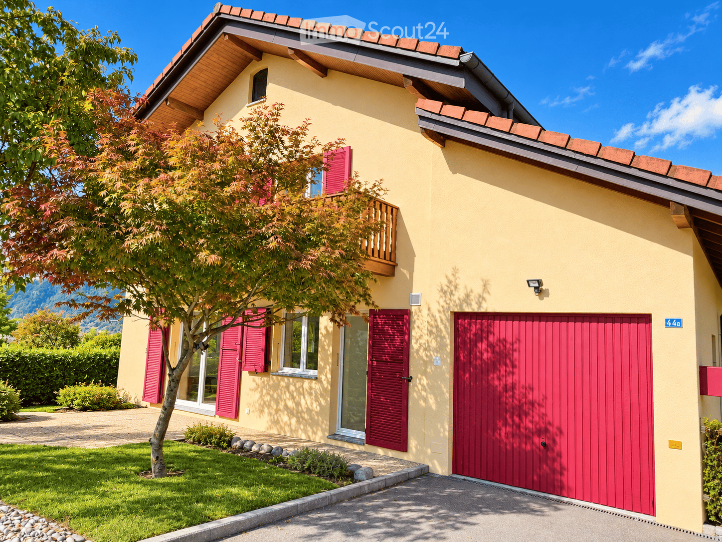 Two-story house with yellow exterior, red shutters, garage, and balcony overlooking a garden area.