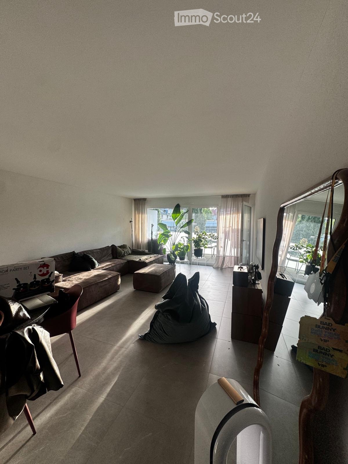 Modern living room with a large sectional sofa, ottoman, beanie bag chair, and a wooden side table with a mirror. The room features large windows with curtains and several potted plants, creating a bright and inviting atmosphere.