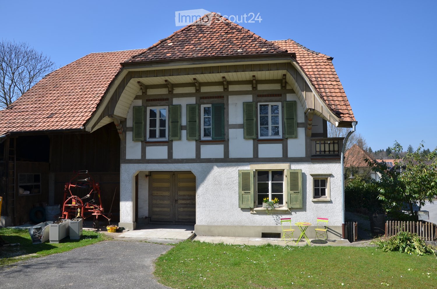 Two-story traditional house with a tiled roof, white and brown facade, green shutters, and a small garden area with chairs and a table.