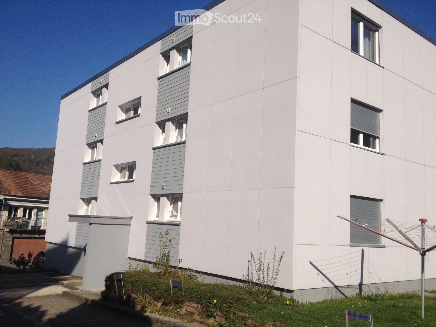 Modern multi-story building with white facade, grey accents, and multiple windows with shutters. Small garden area in front with some plants and a clothesline.