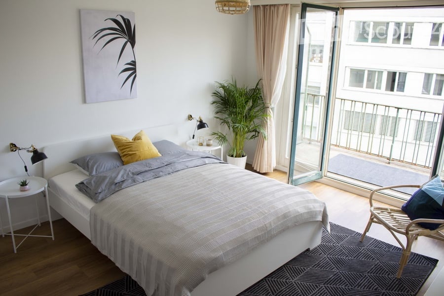 Modern bedroom with a white bed, grey bedding, nightstand, chair, and a large window opening to a balcony.