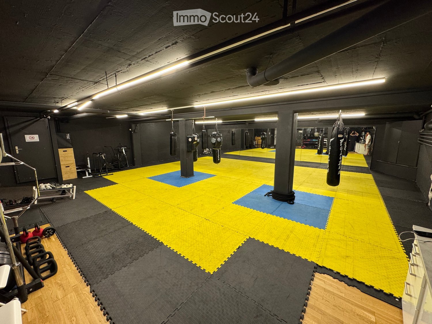 A spacious gym area with a large yellow and blue mat, boxing bags hanging from the ceiling, weights, a bench, and a mirror on the wall.