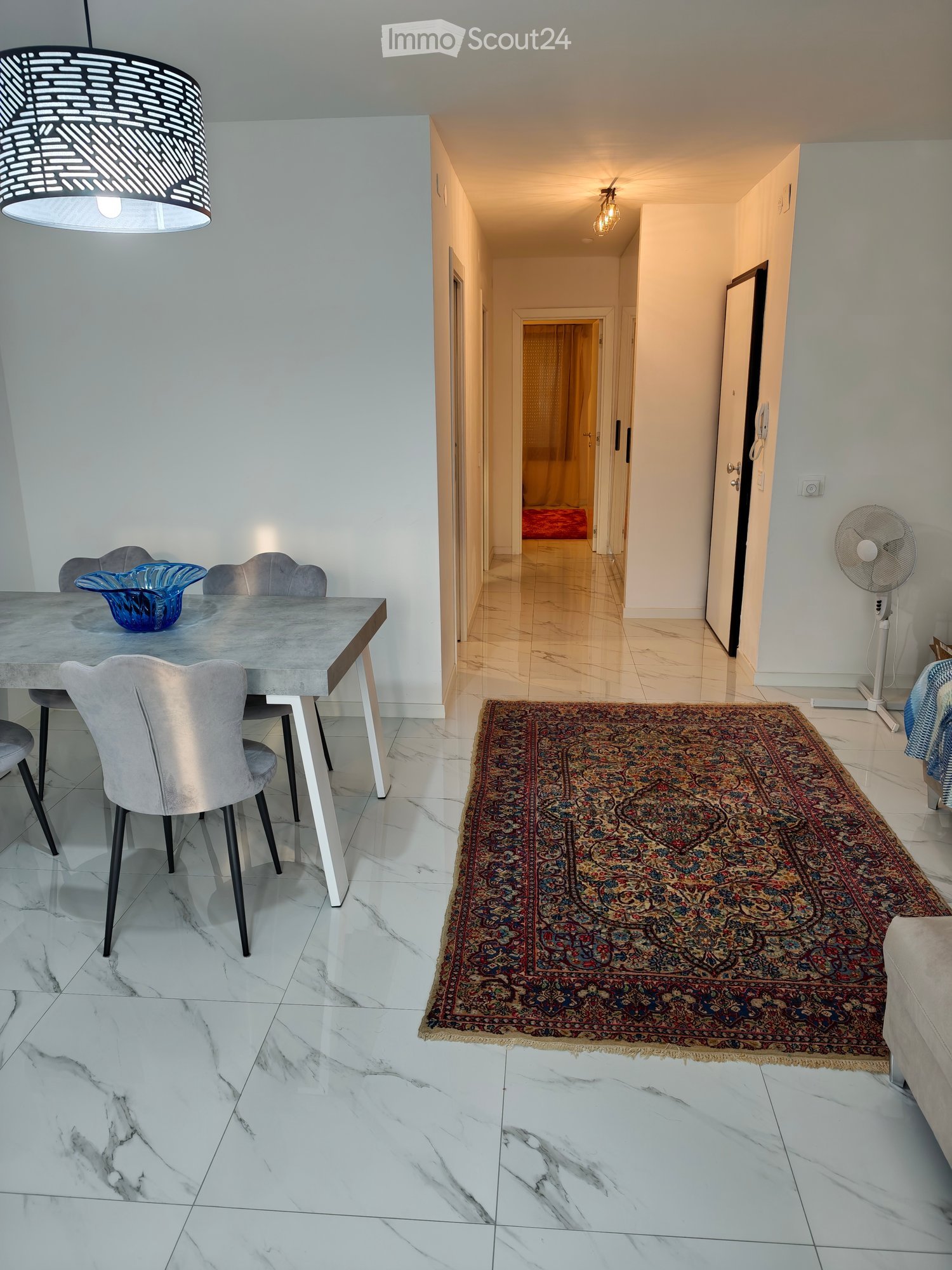 Modern hallway with marble flooring, a patterned rug, and a dining area visible to the left, featuring a grey table, chairs, and a decorative bowl.