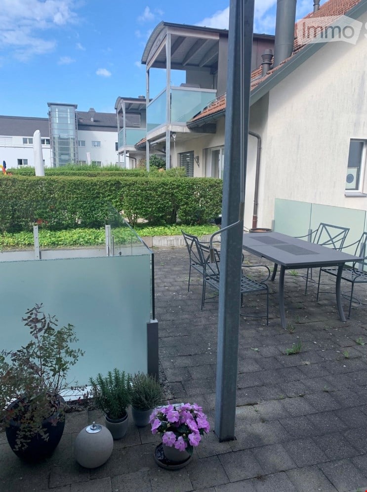 Balcony area with a table, chairs, and potted plants, surrounded by modern buildings and greenery.