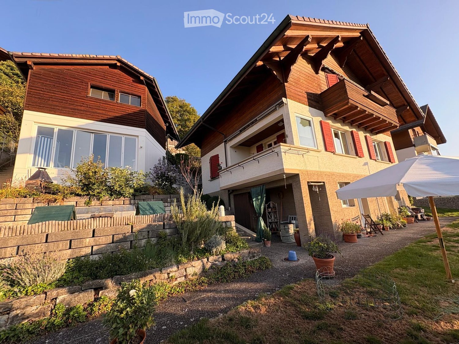 Two-story house with wooden exterior, red shutters, and a balcony. The garden features potted plants, a stone wall, and a patio area with outdoor furniture.