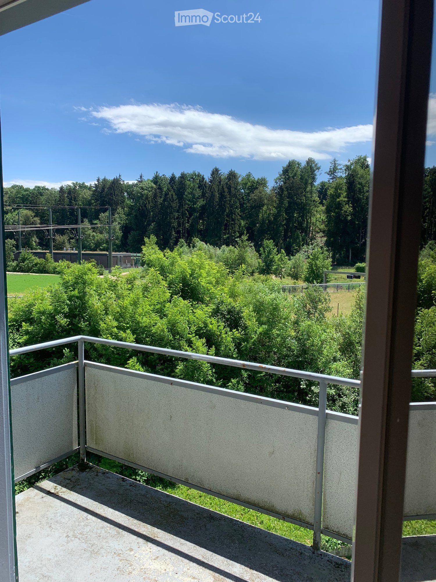 Balcony with metal railing, overlooking a lush green landscape with trees and a clear blue sky.
