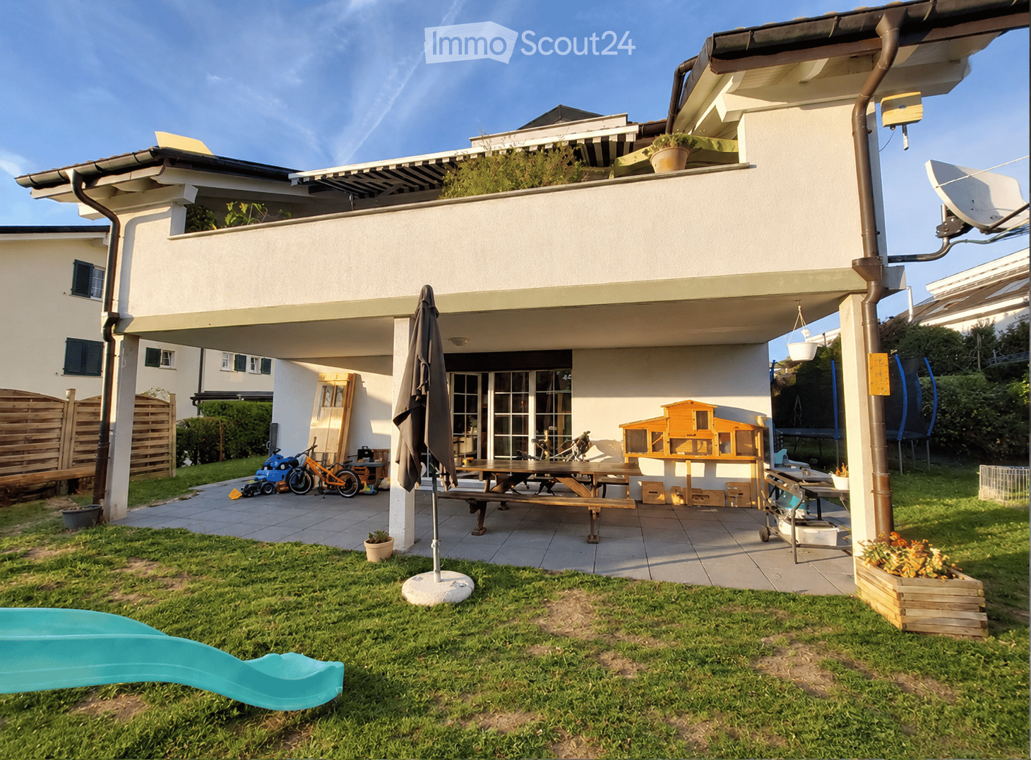 A well-maintained garden area with a patio, picnic table, bicycles, and a small play area, featuring a modern house with a raised ground floor and a covered outdoor space.