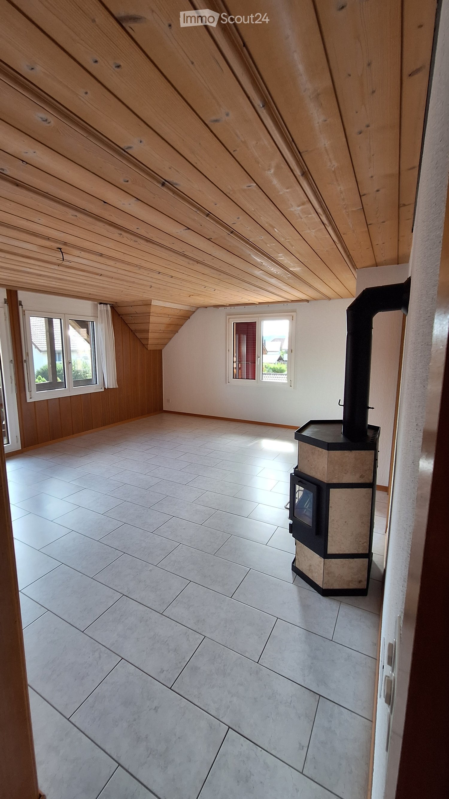 Empty attic room with wooden slanted ceiling, tiled floor, and a fireplace in the corner. Two windows provide natural light.