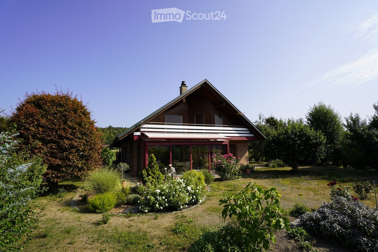 Two-story house with a wooden balcony, surrounded by a well-maintained garden with various plants and trees.