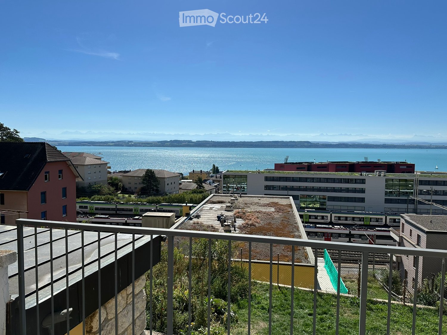 Balcony overlooking a scenic view of a lake, distant mountains, and a town with buildings and train tracks.