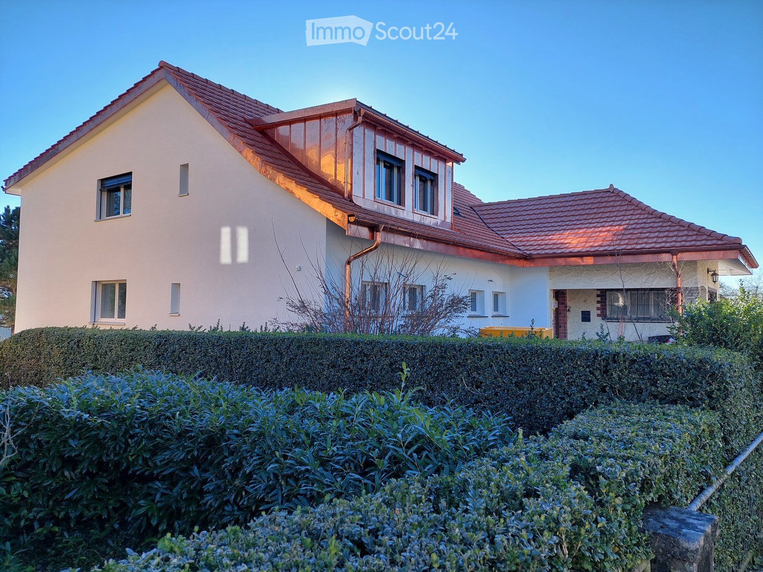 Two-story house with red-tiled roof, white walls, multiple windows, and a well-maintained hedge in the front yard.