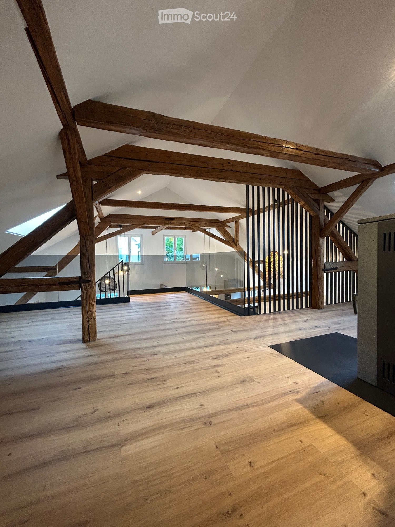Open, airy space with exposed wooden beams and a polished wooden floor, featuring a modern glass railing and large windows allowing natural light.