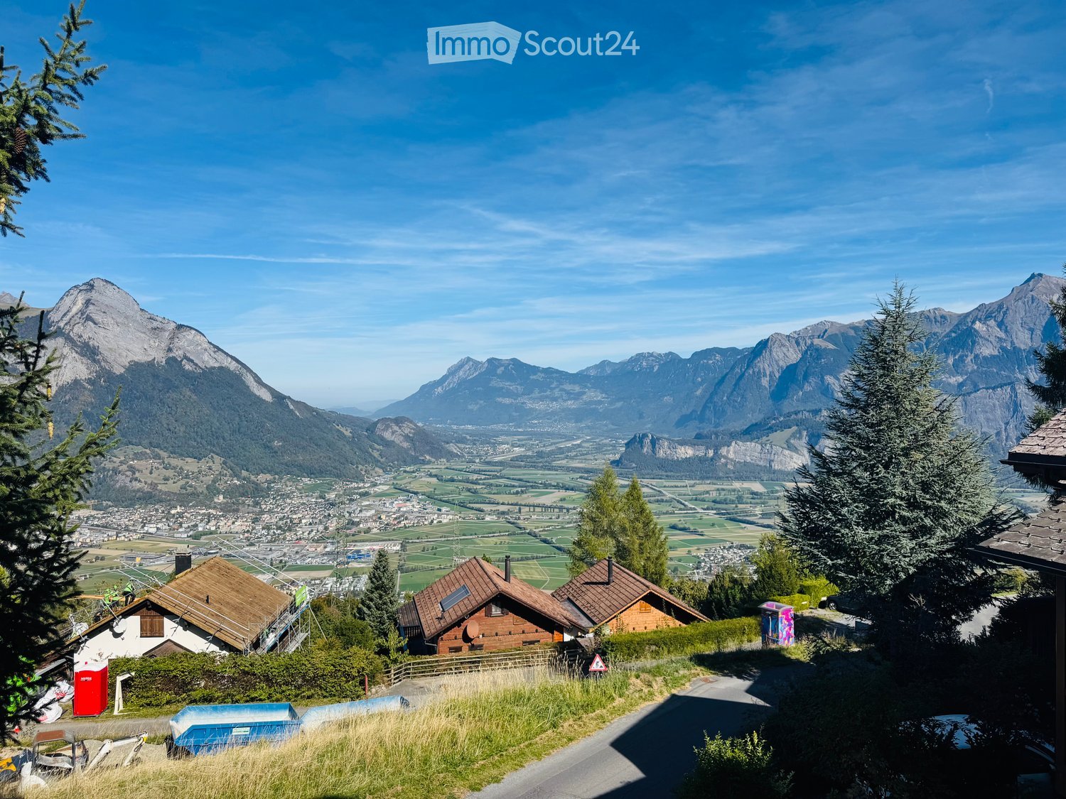 A scenic mountain view with a town below, featuring several houses with wooden roofs and surrounding greenery.