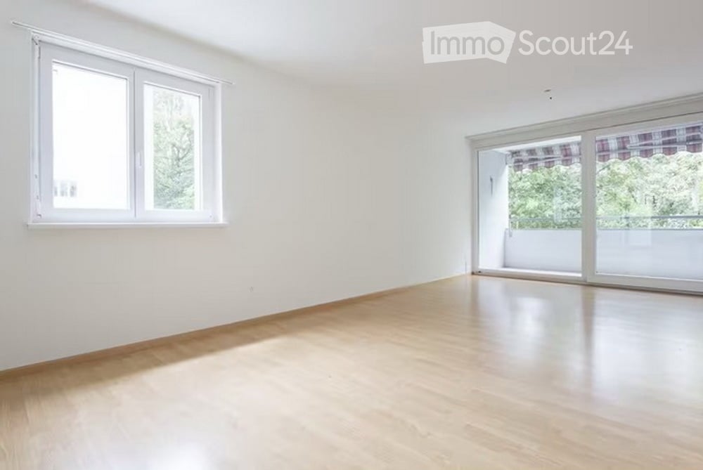 Empty room with light wooden flooring, white walls, large windows, and a sliding glass door leading to a balcony.