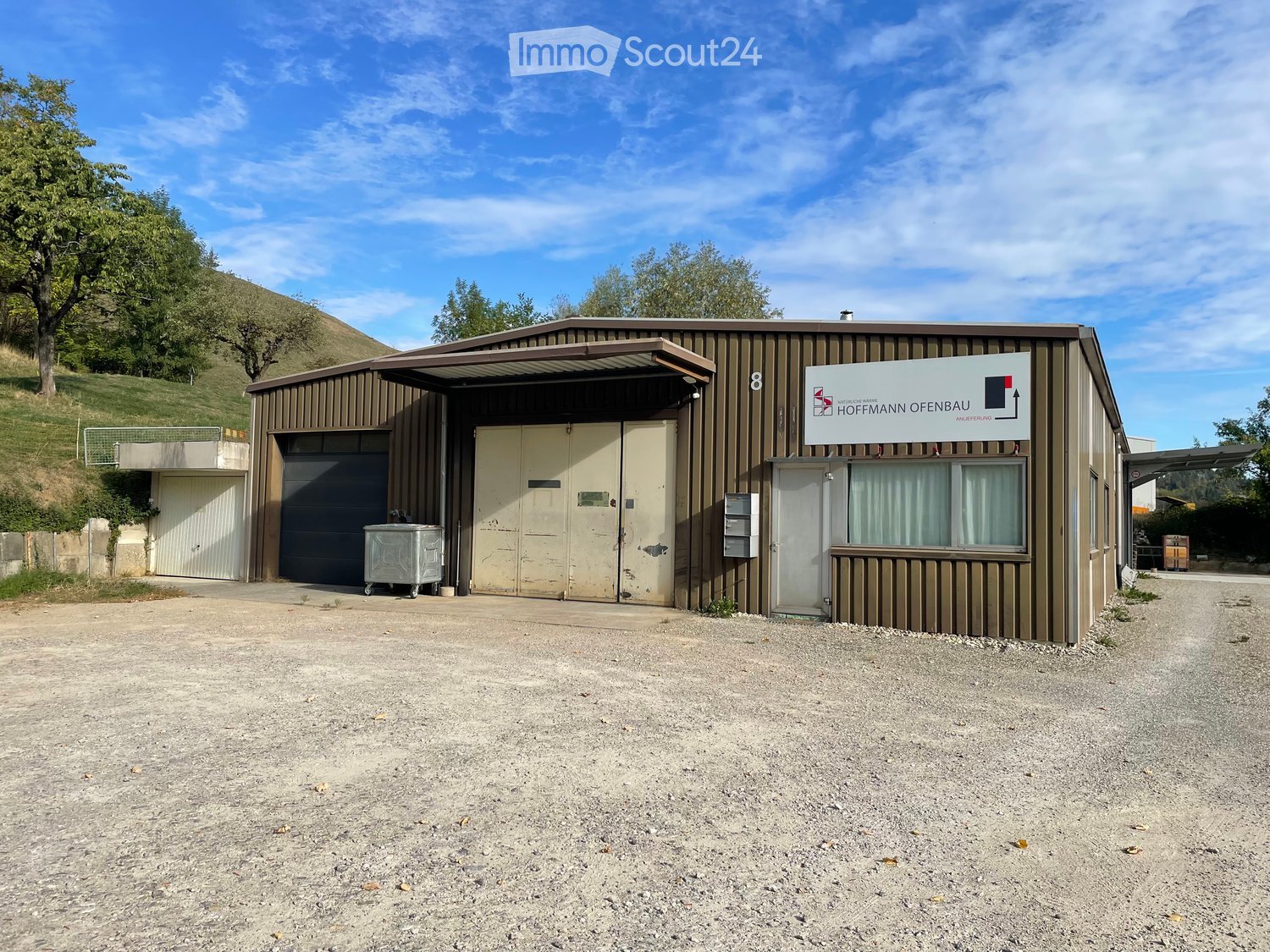 A modern industrial-style building with a large garage door, a sign reading "NATURLICHE WARME HOFFMANN OFENBAU," and a small windowed section. The building is situated on a gravel surface with a grassy hill in the background.