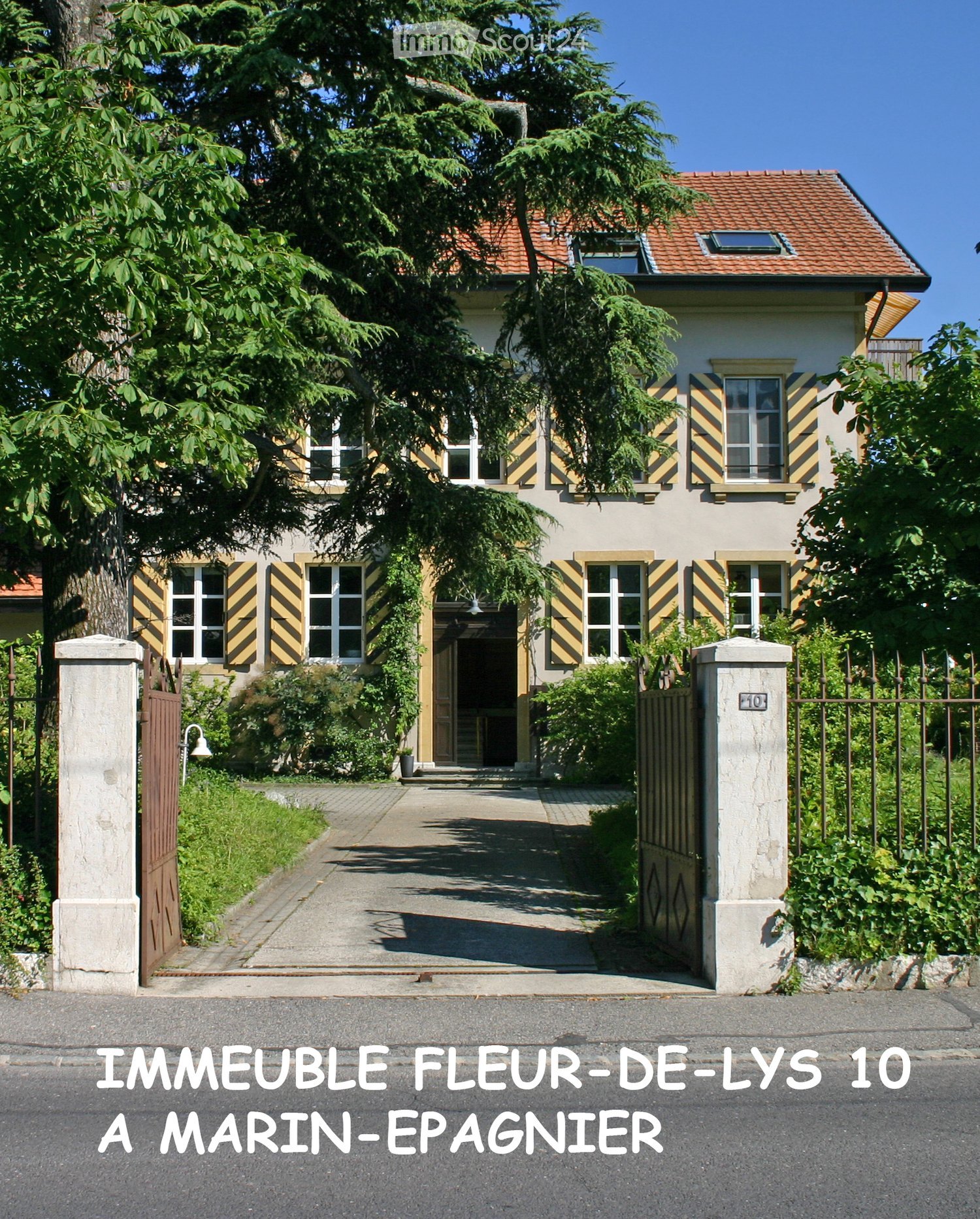 Two-story house with a red-tiled roof, white walls, and yellow shutters. The entrance features a small porch with steps leading up to a wooden door. The house is surrounded by greenery and has a gated driveway.