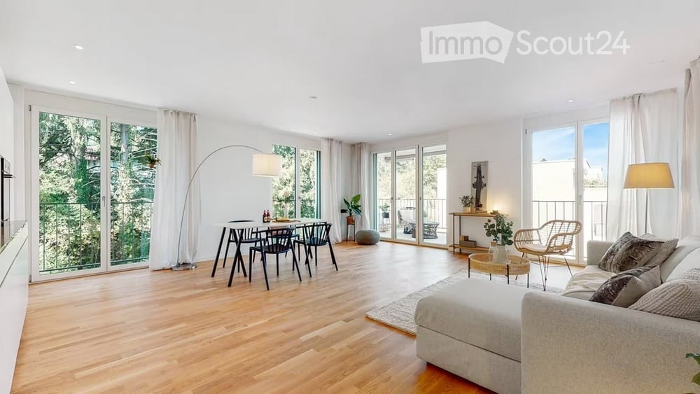 Bright, open-plan living and dining area with wooden flooring, large windows, and sliding glass doors leading to a balcony. Features a white sofa, coffee table, dining table with chairs, floor lamp, and side table with decor.