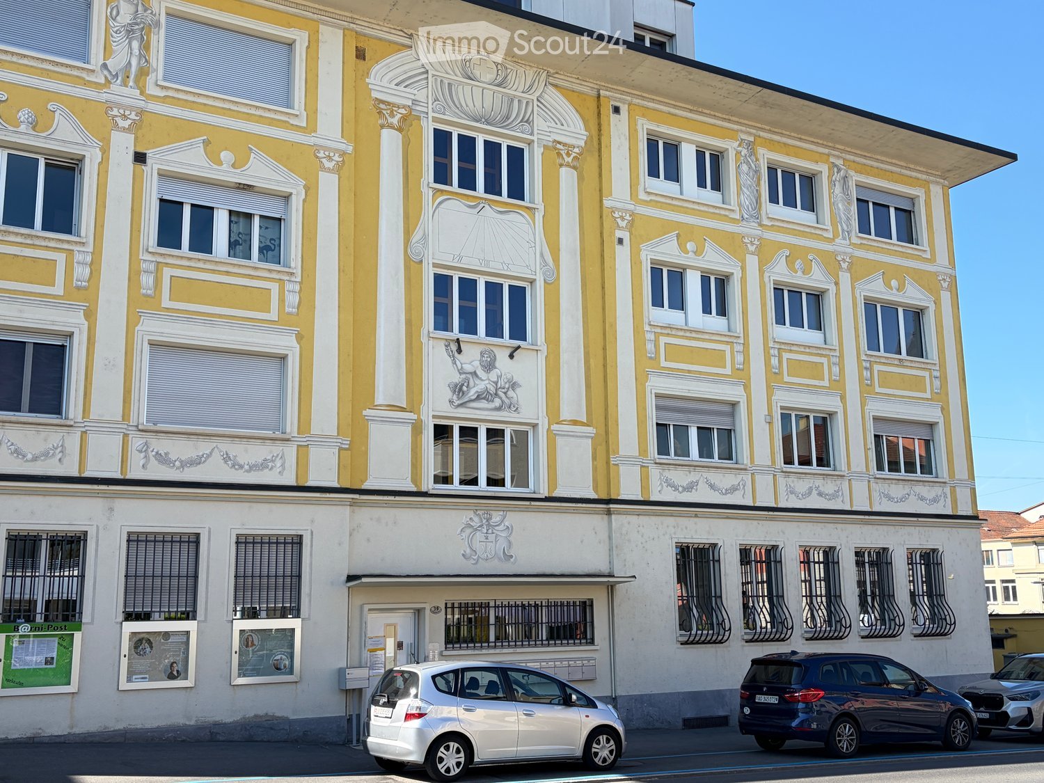 A multi-story building with yellow and white facade, decorative columns, and ornate carvings. Windows have shutters, and there is a lower section with a door and posters.