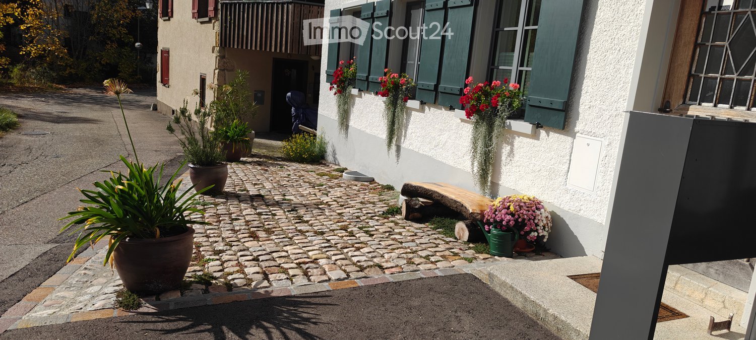 A cobblestone pathway leads to a house entrance, flanked by potted plants and hanging flower baskets. The house features white walls, green shutters, and a small wooden bench near the entrance.