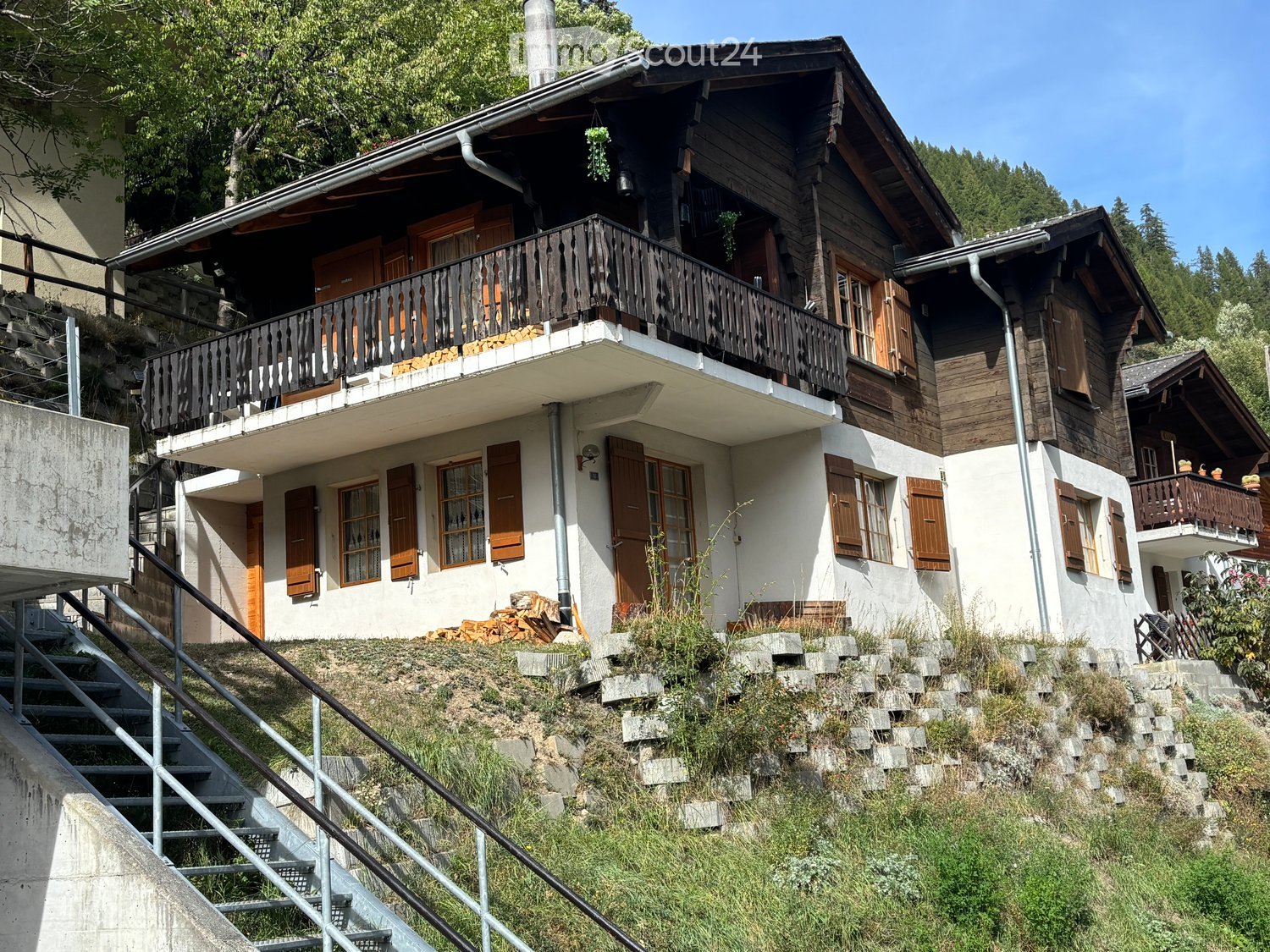 A two-story wooden house with white walls, wooden shutters, and balconies, situated on a raised ground floor with stone steps leading up to it.