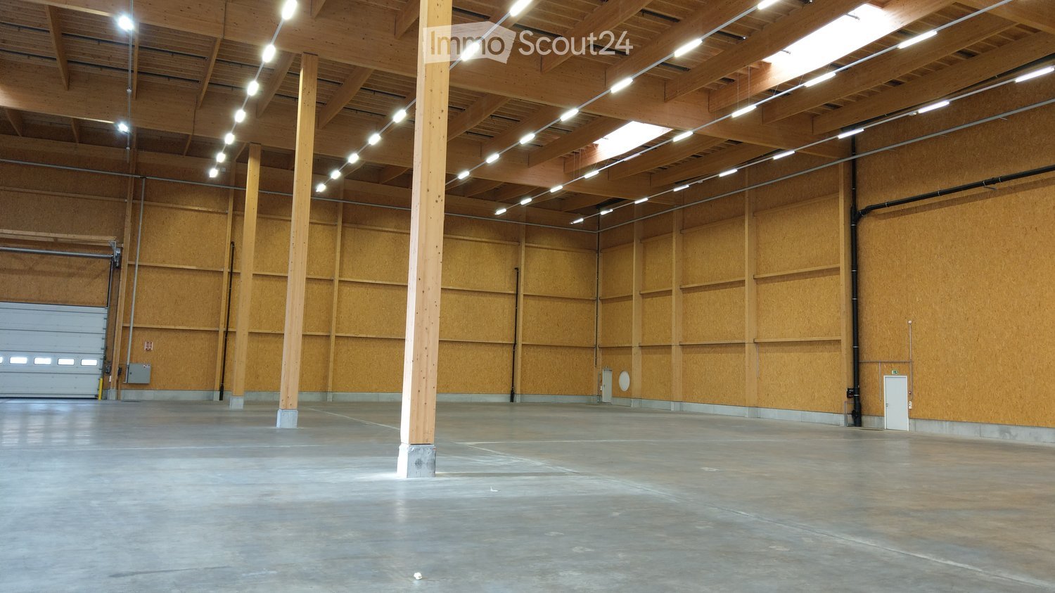 Empty warehouse space with wooden walls, concrete floor, exposed wooden beams, and a large garage door.
