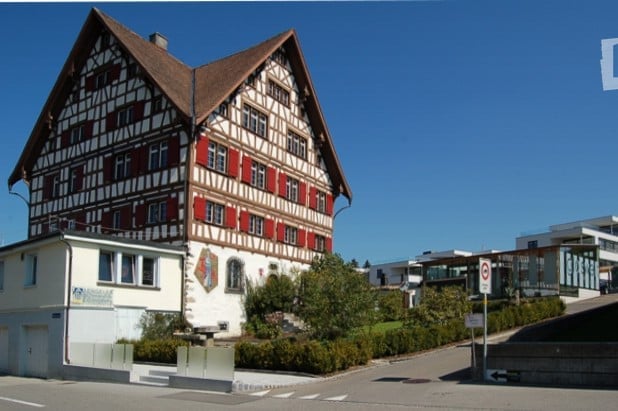 Traditional Swiss-style building with a steeply pitched roof, exposed wooden beams, and red shutters. The structure is two stories with multiple windows and a small balcony on the upper level.