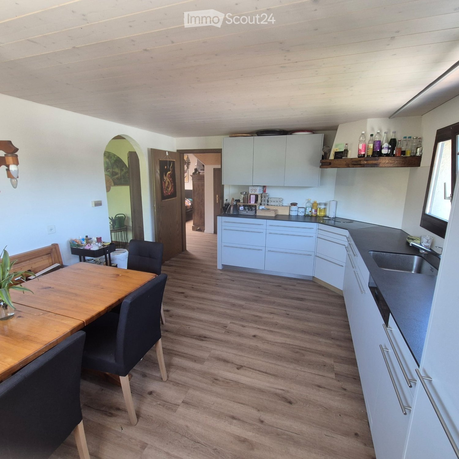 A modern kitchen with light grey cabinets, a wooden dining table with chairs, and a sink area with various bottles and cups on the counter.