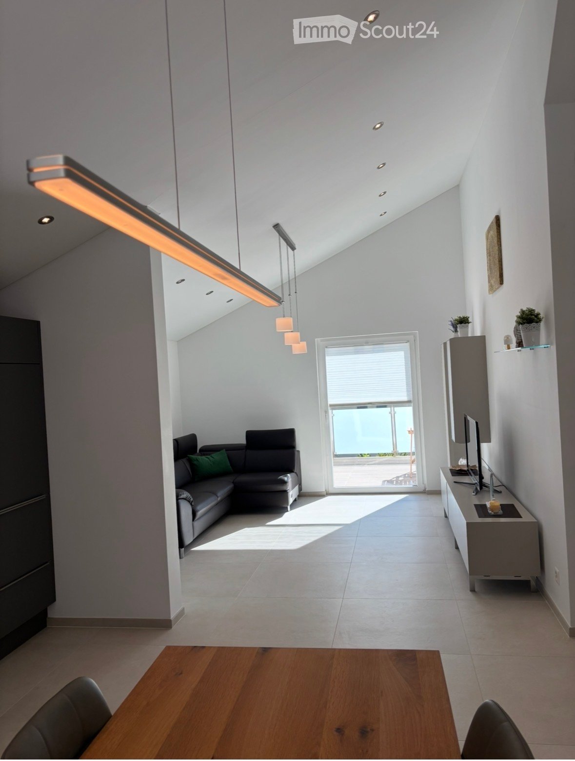 Modern living room with a grey sectional sofa, wooden dining table, chairs, TV cabinet, and a view through large windows.