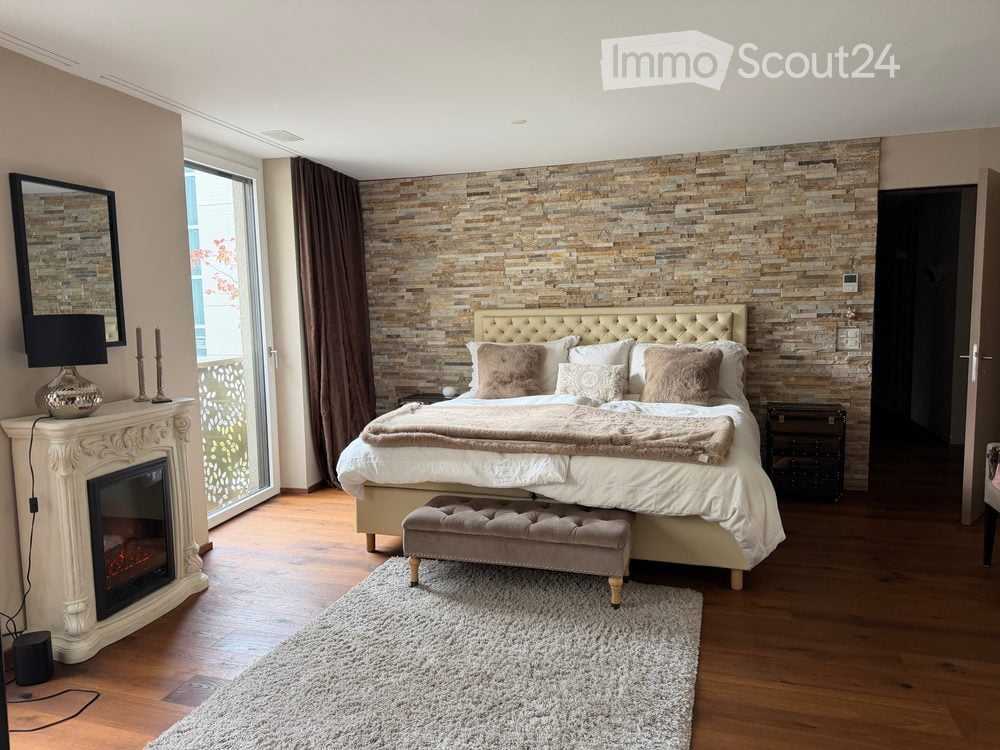 Elegant bedroom with a large bed, fireplace, mirror, and decorative lamp. Features a stone accent wall and large window with curtains.