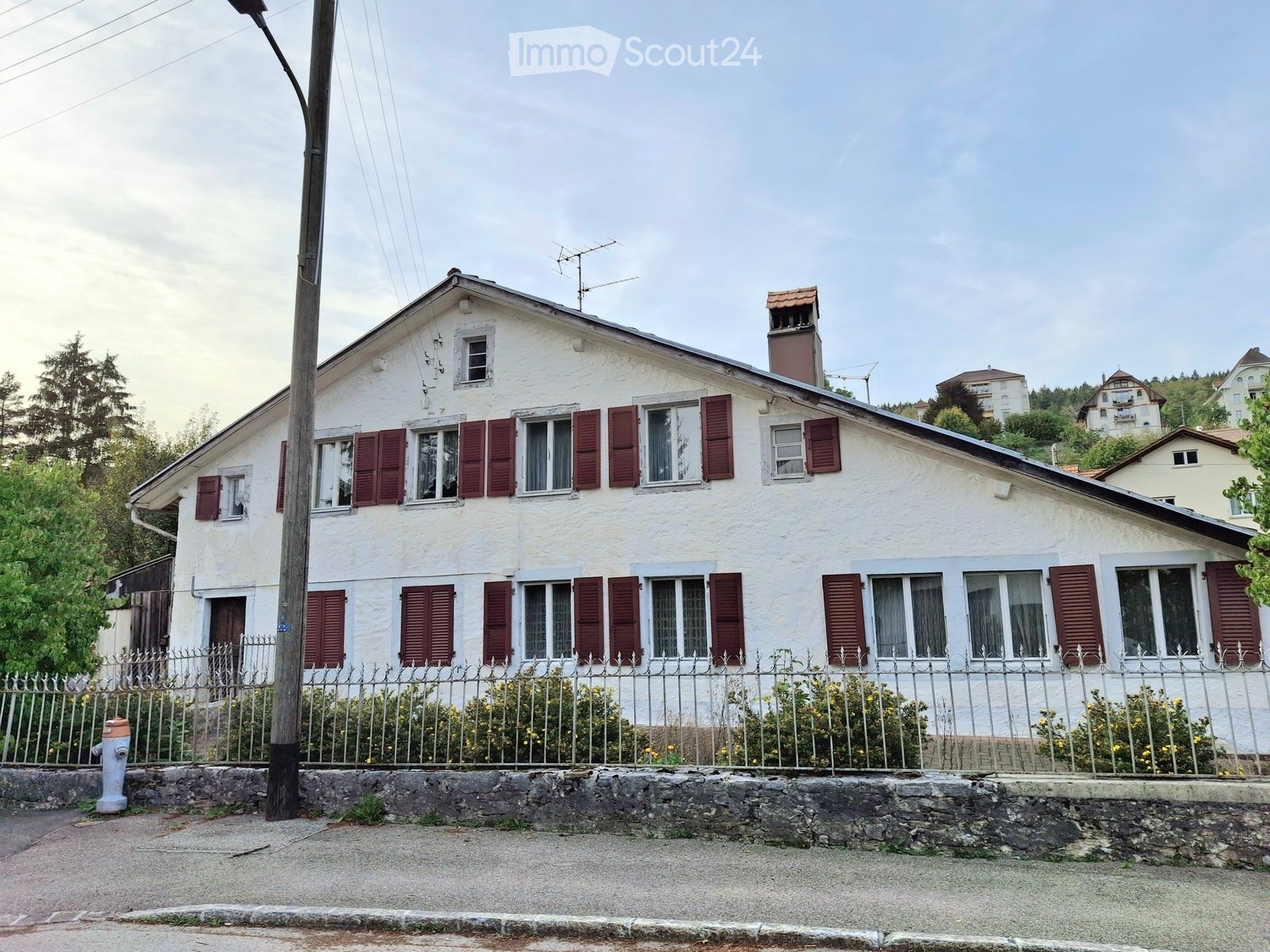 Two-story white house with red shutters, multiple windows, chimney, and a small garden behind a metal fence.