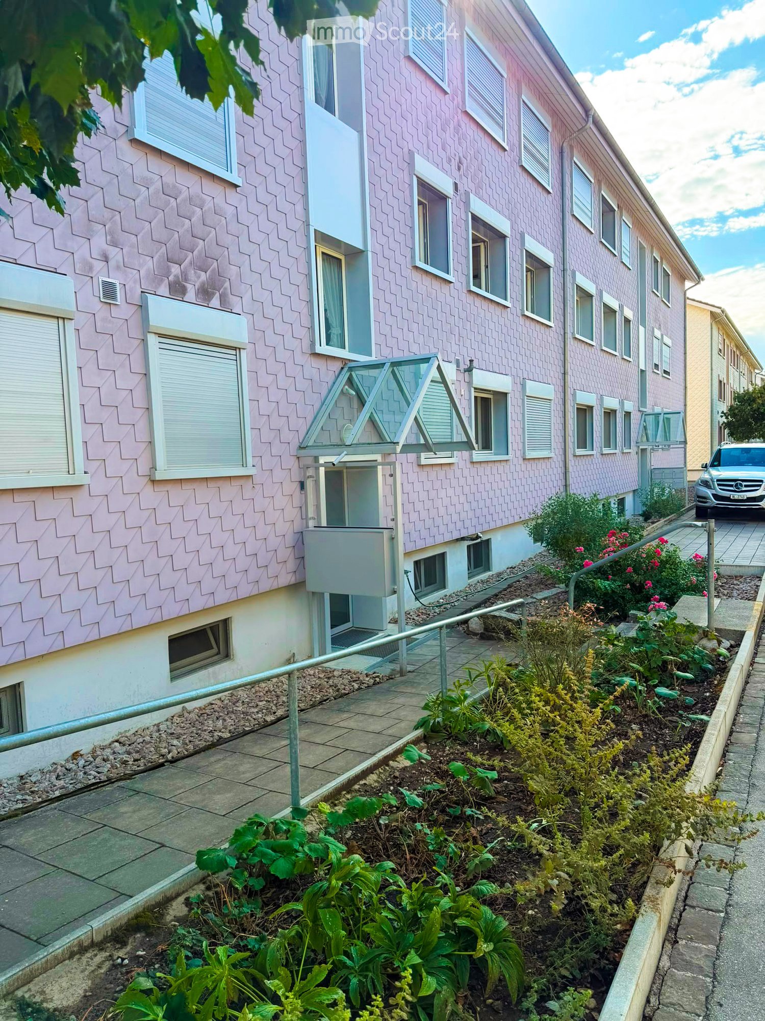 Pink residential building with multiple windows, a small garden area with plants and flowers, and a parked car in the background.