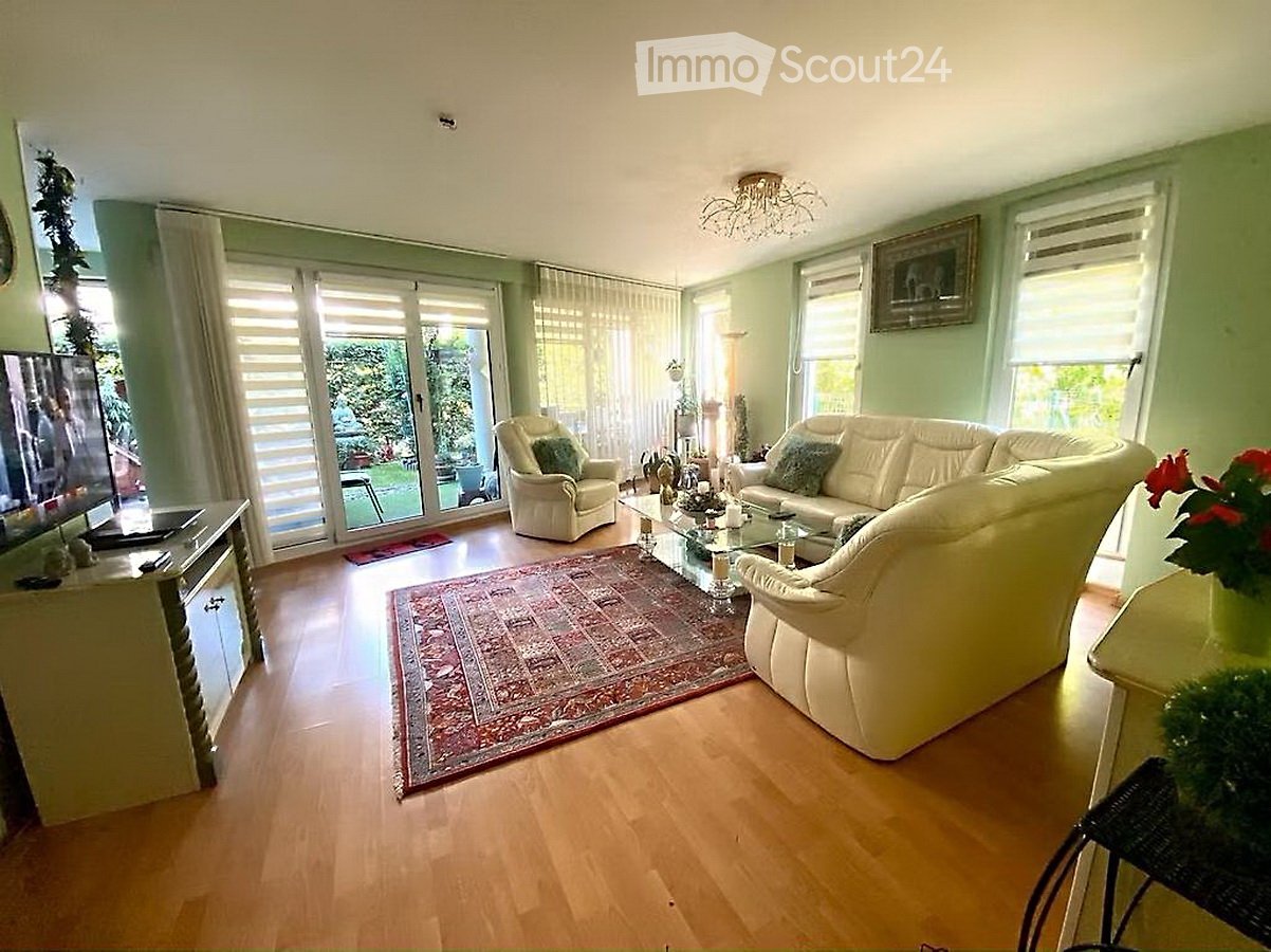 Bright living room with light green walls, wooden flooring, large windows, and a sliding glass door leading to a garden. Features a cream-colored sofa, armchair, coffee table, television, and a decorative rug.