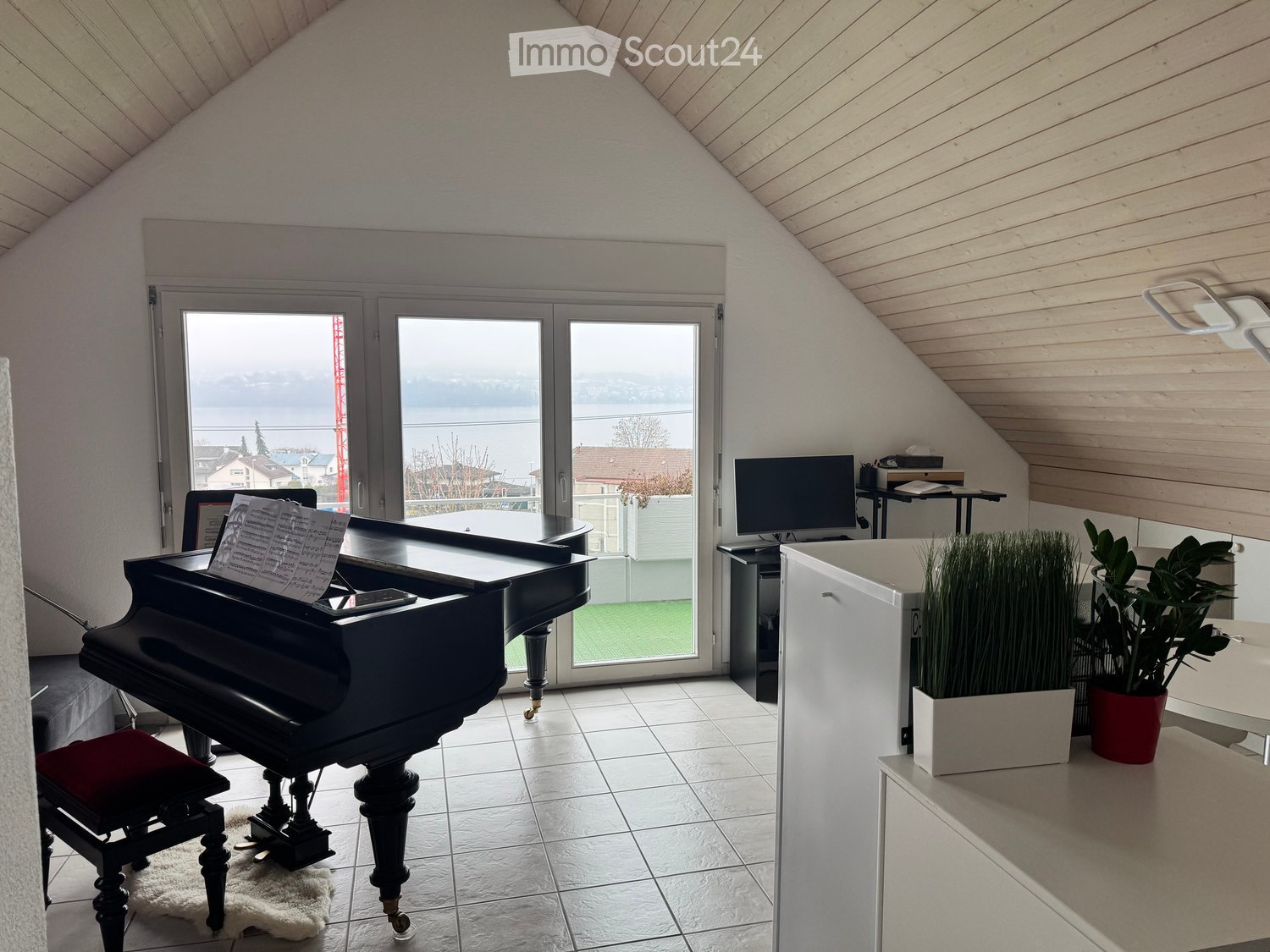 Bright room with a grand piano, a red stool, a television, a table, and potted plants, offering a scenic view through large windows.