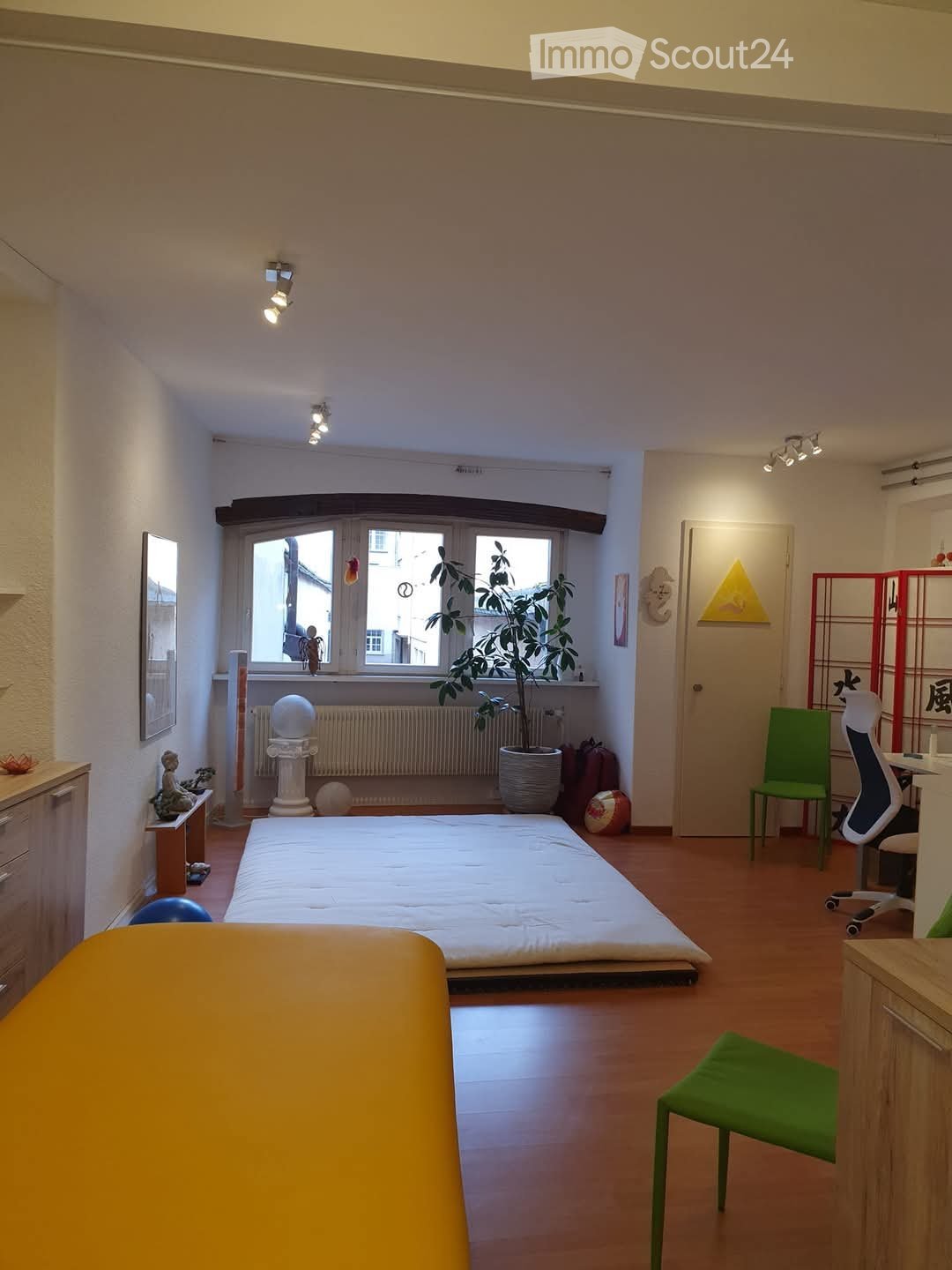 A well-lit bedroom with a tatami-style bed, a green chair, a desk with a chair, a potted plant, a mirror, and a small cabinet.