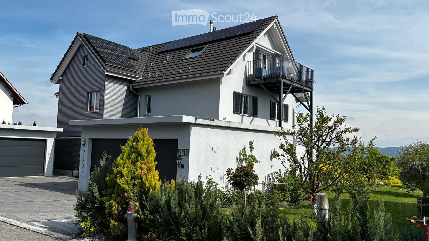 Modern two-story house with grey siding, solar panels on the roof, a balcony on the upper level, and a garage with a driveway.