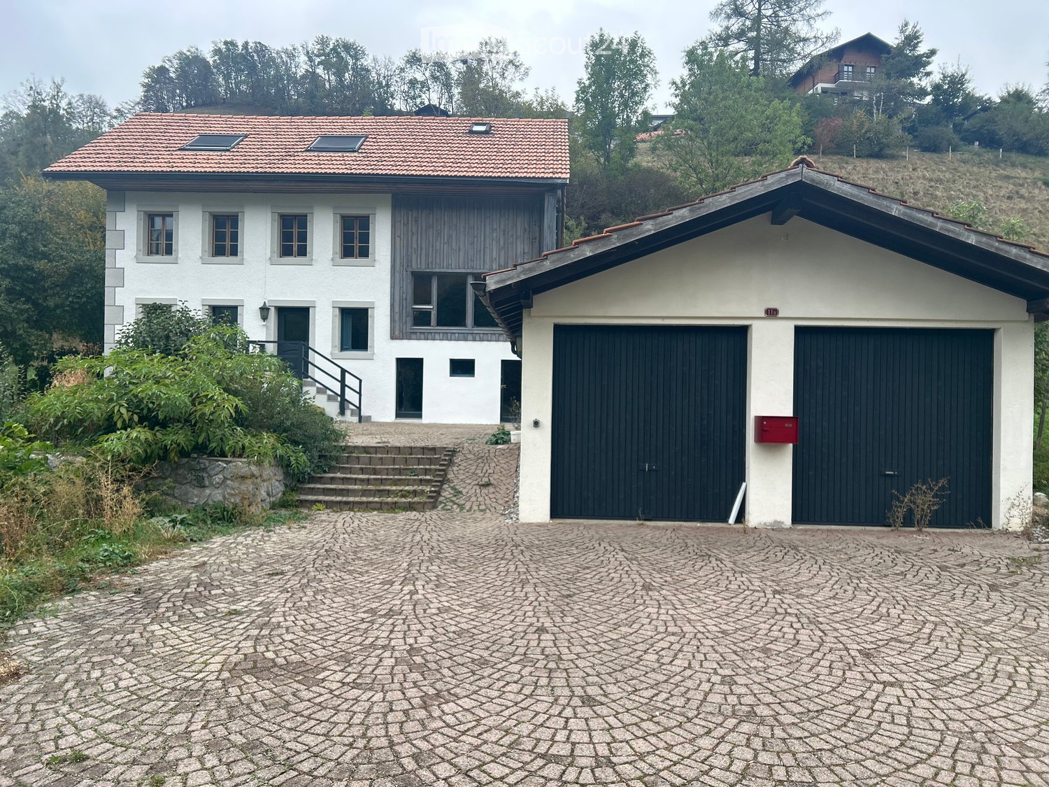 Two-story house with white exterior, red-tiled roof, and attached garage with black doors. The house features multiple windows and a small staircase leading to the entrance.