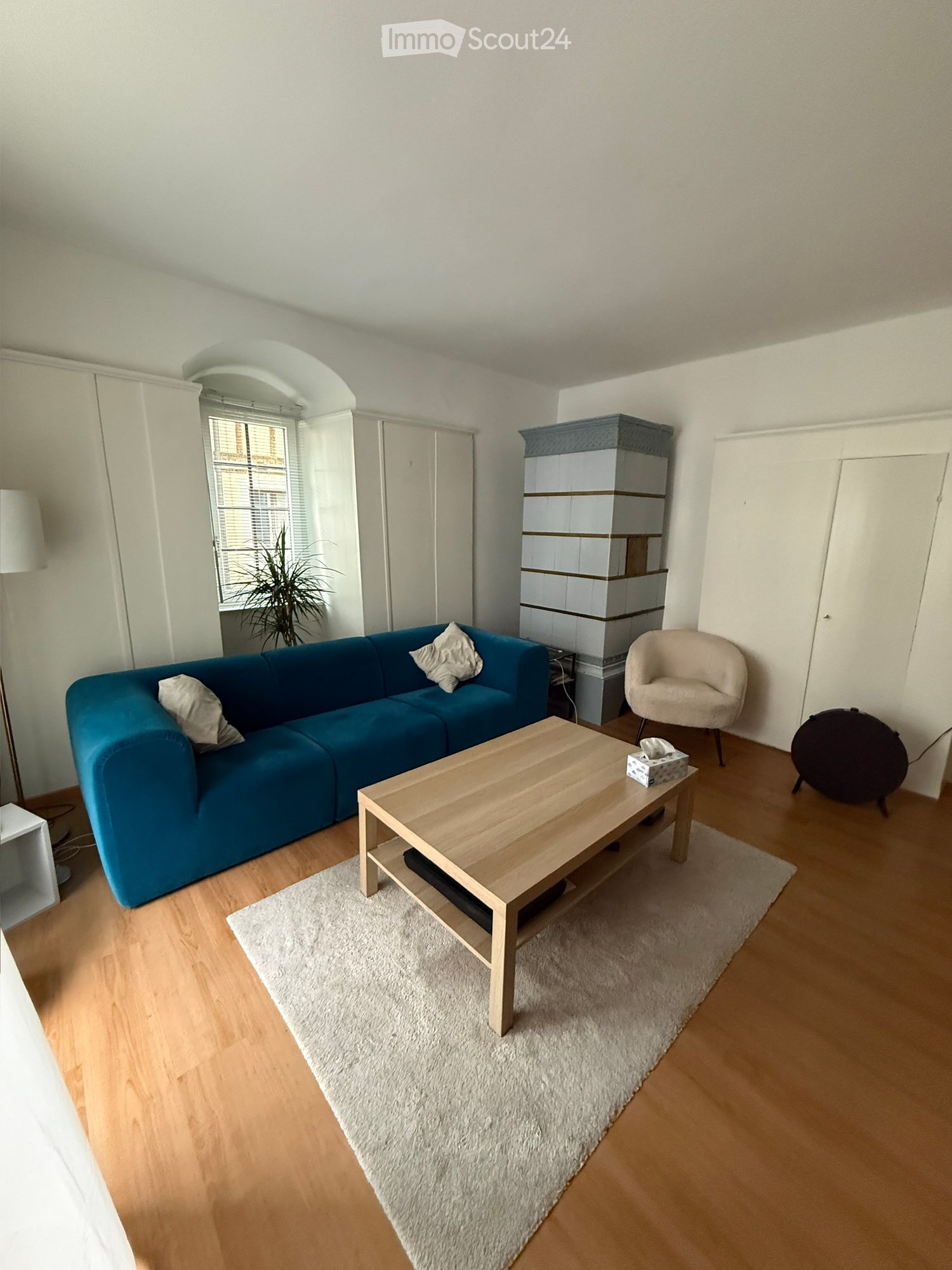 Bright living room with a blue sofa, wooden coffee table, white shaggy rug, and a small round chair. Features a large window with blinds and a storage unit in the corner.