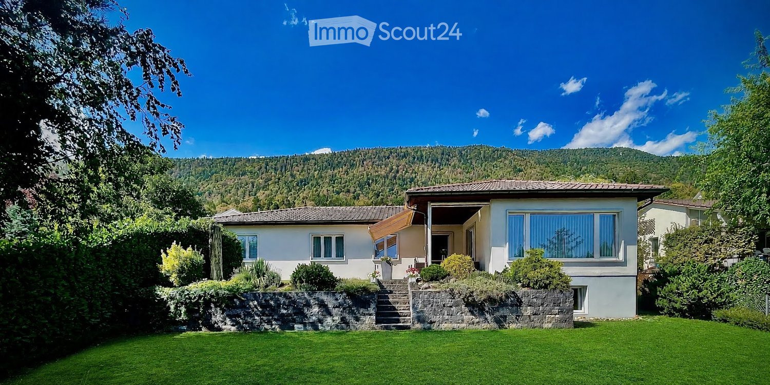 Single-story house with white walls, multiple windows, and a tiled roof, surrounded by a well-maintained lawn and garden, with a scenic mountain backdrop.