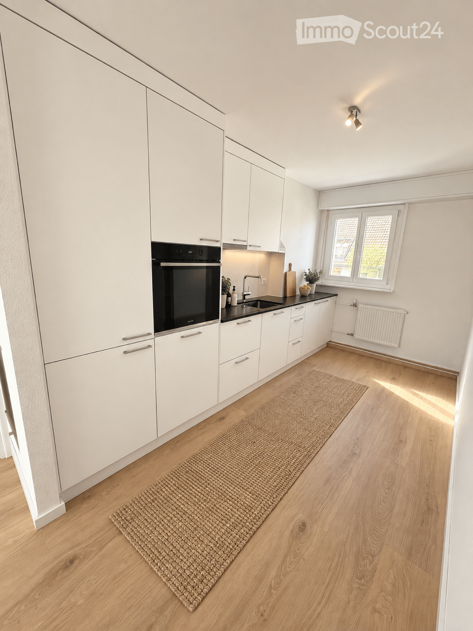 Modern kitchen with white cabinets, stainless steel oven, sink, and faucet. Light wooden flooring and a woven rug in the center.
