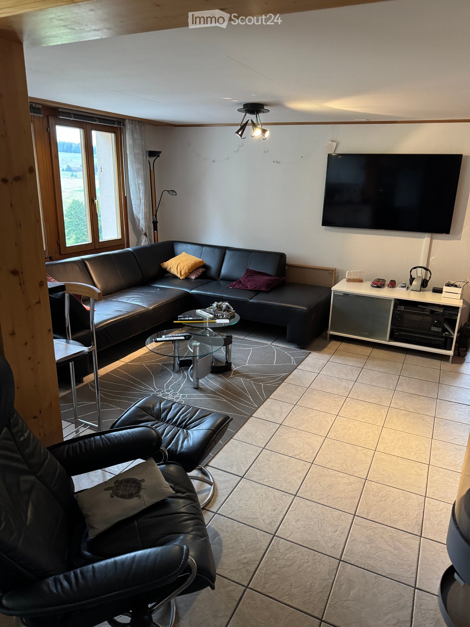 Modern living room with a black leather sectional sofa, a black leather armchair, a glass coffee table, a flat-screen TV, and a small white table with various items on it.